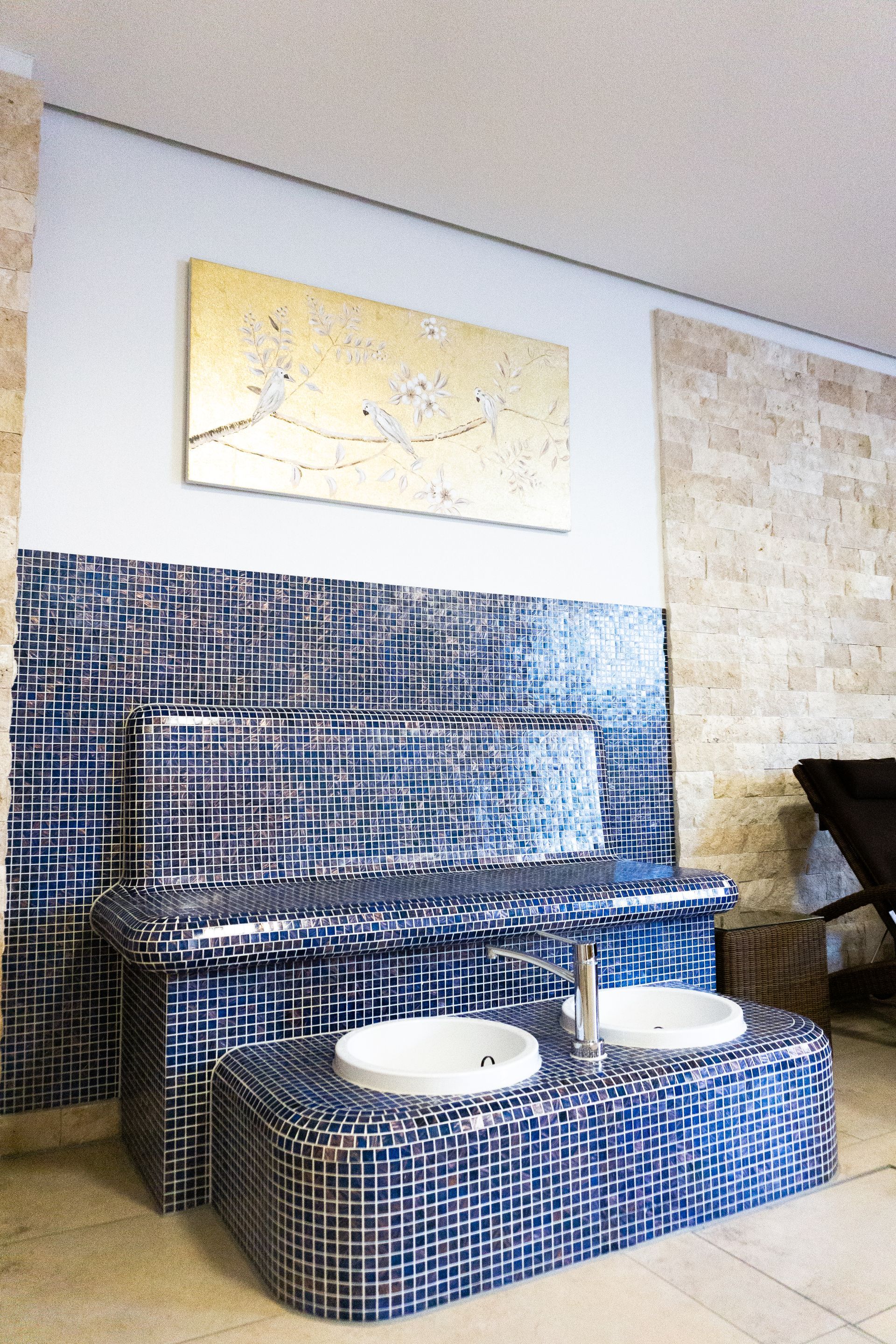 Blau geflieste Badezimmerwaschbecken mit weißen Becken vor einer Mosaikwand und goldenen Kunstwerken.