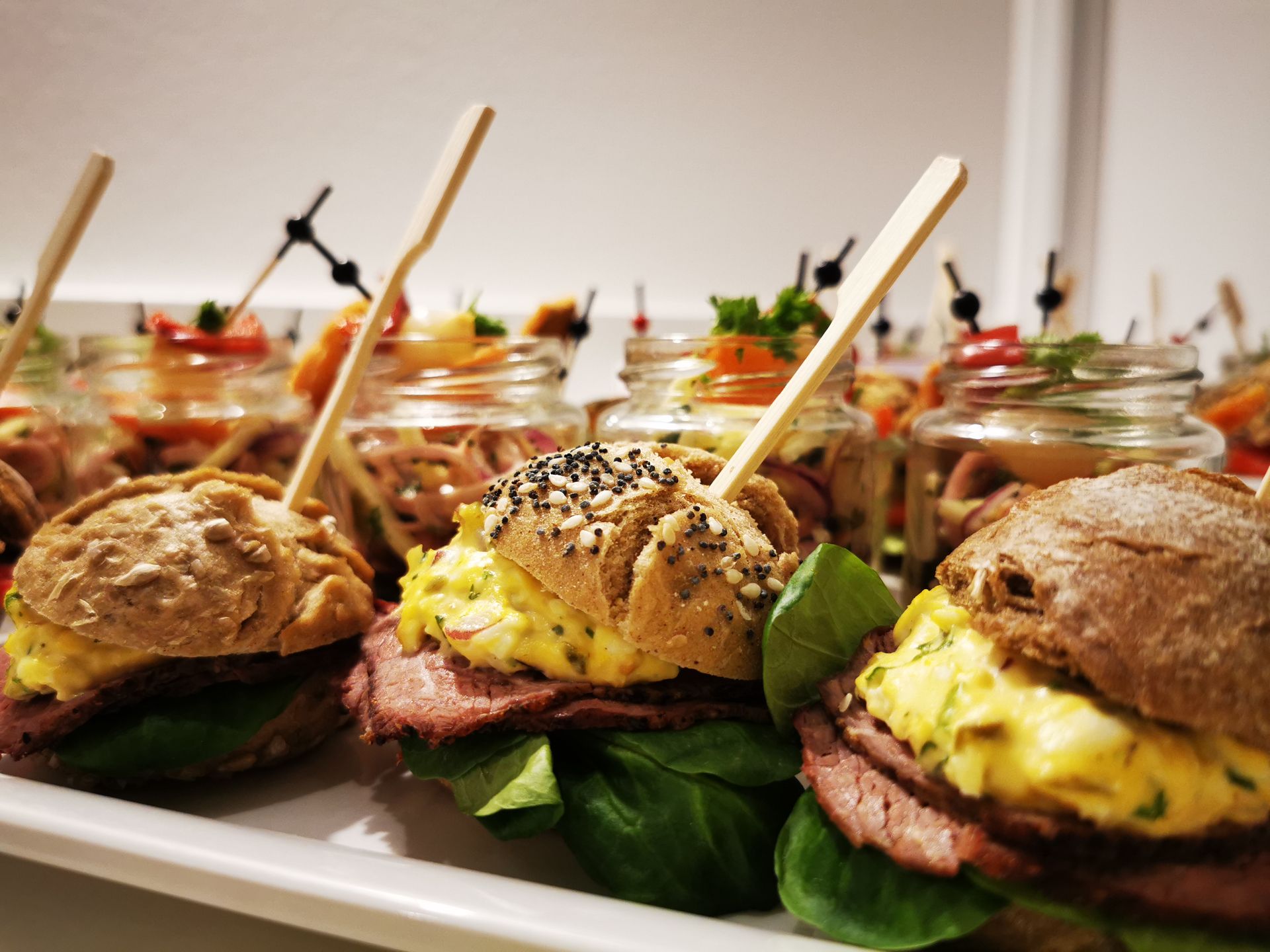 Mini-Sandwiches mit Rindfleisch und Eiersalat auf einer Platte. Gläser mit Salat im Hintergrund.