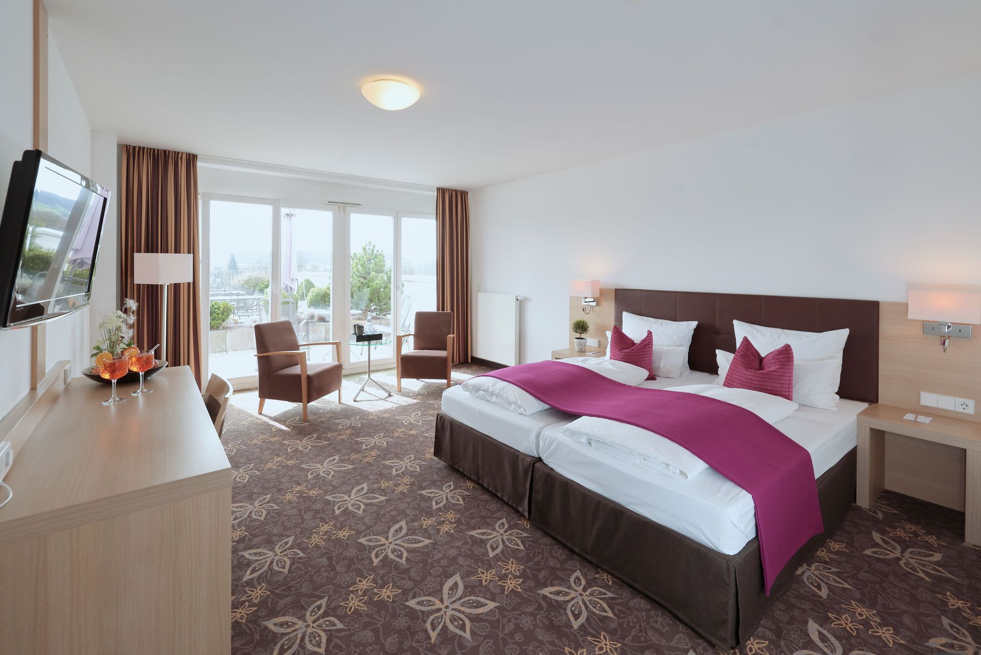 Hotelzimmer mit Doppelbett, Schreibtisch und Balkon. Beige Wände, braune Vorhänge und geblümter Teppich.