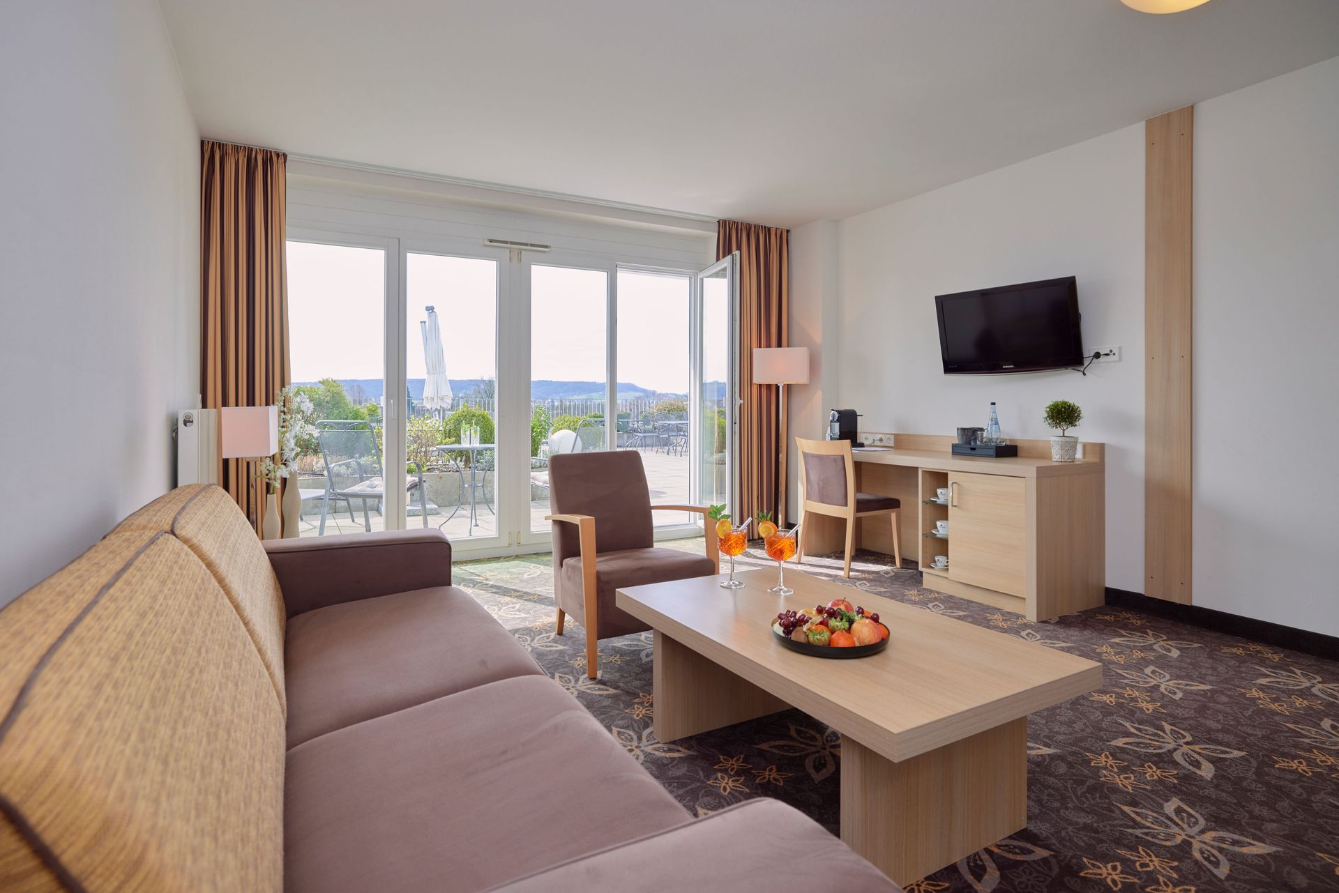 Wohnzimmer mit Sofa, Sessel, Schreibtisch, TV und Zugang zur Terrasse, Blick auf einen Balkon mit Gartenmöbeln.