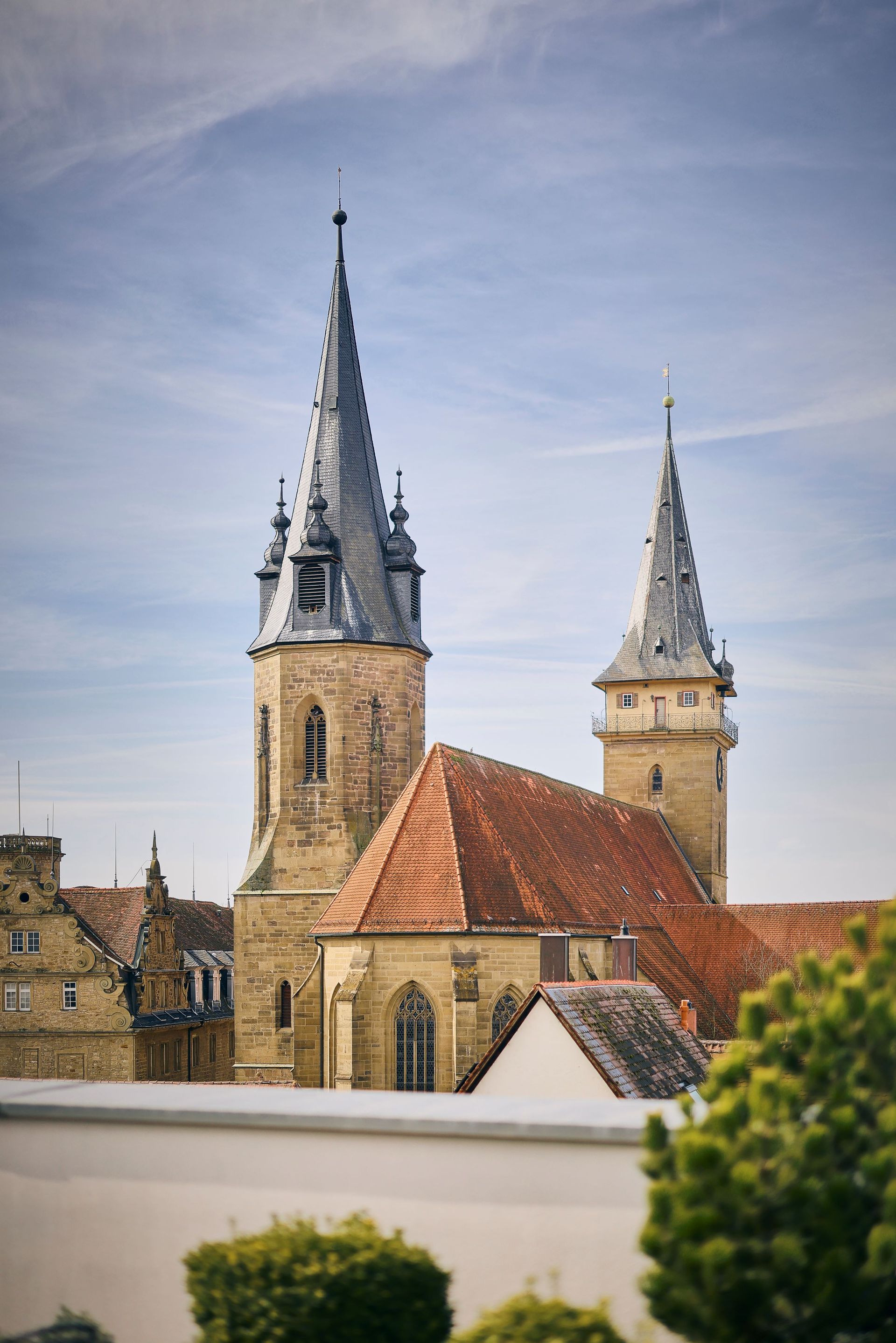 Unter einem bewölkten Himmel erheben sich zwei hohe Kirchtürme über roten Ziegeldächern.