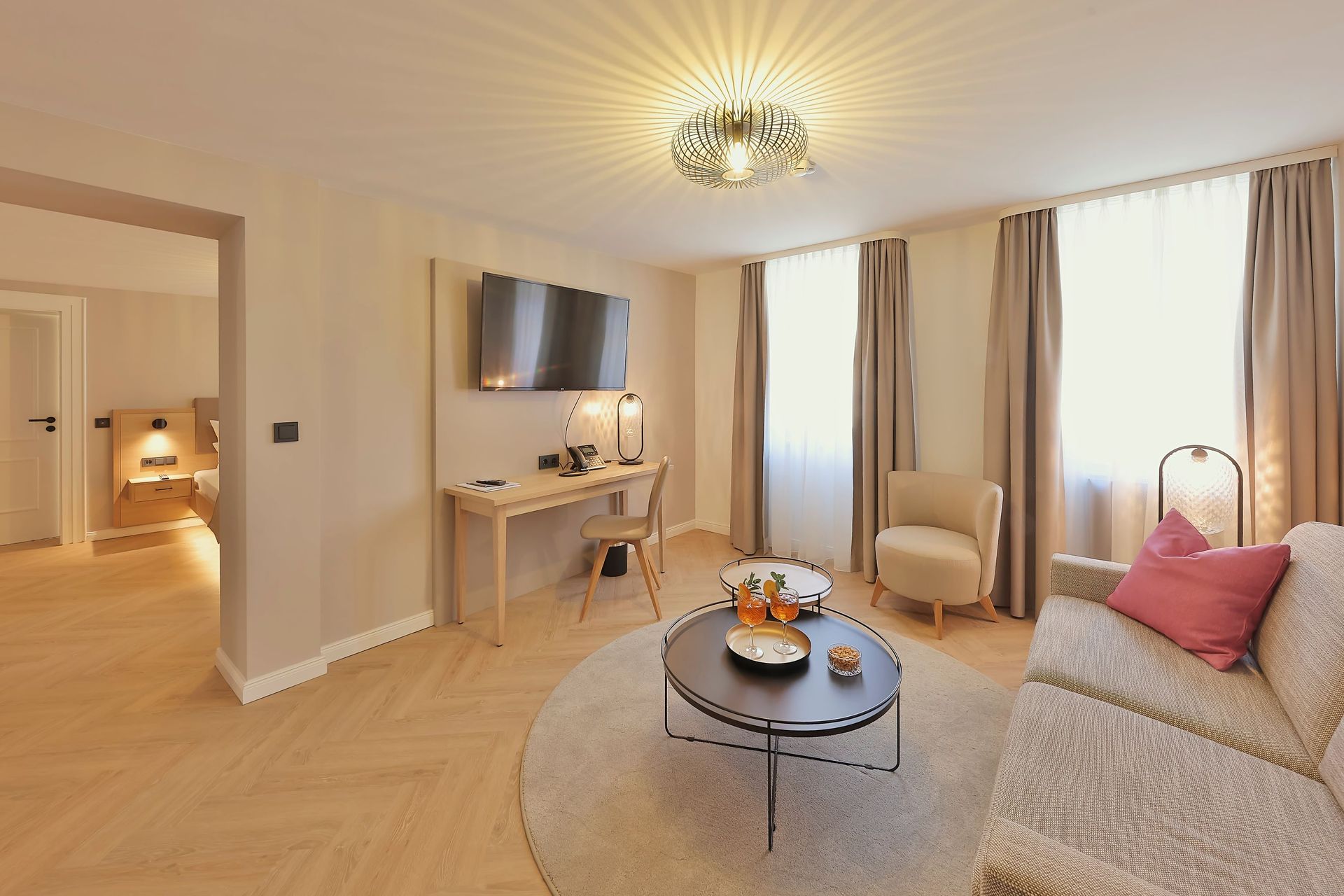 Wohnzimmer mit beigen Wänden, Sofa, Sessel, Schreibtisch und Fenstern mit Vorhängen.