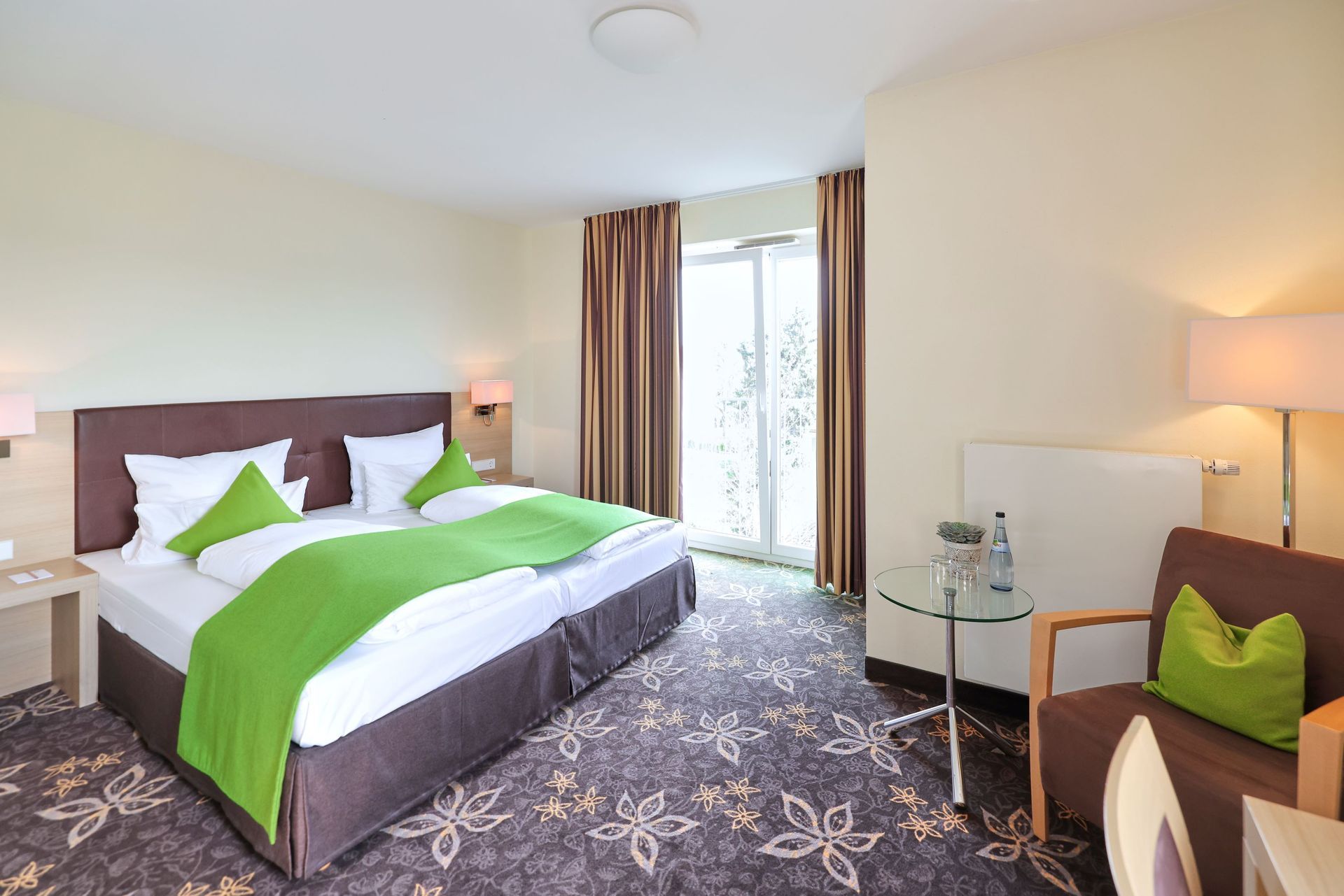 Hotelzimmer mit Doppelbett, Stuhl, kleinem Tisch und Balkon. Farbschema in Grün und Braun.