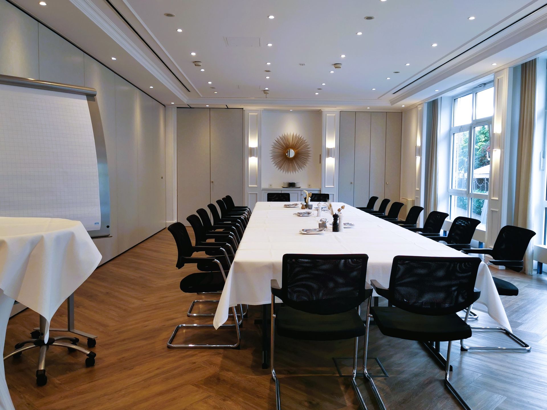 Konferenzraum mit langem Tisch, Stühlen und Flipchart. Natürliches Licht strömt durch ein großes Fenster herein.