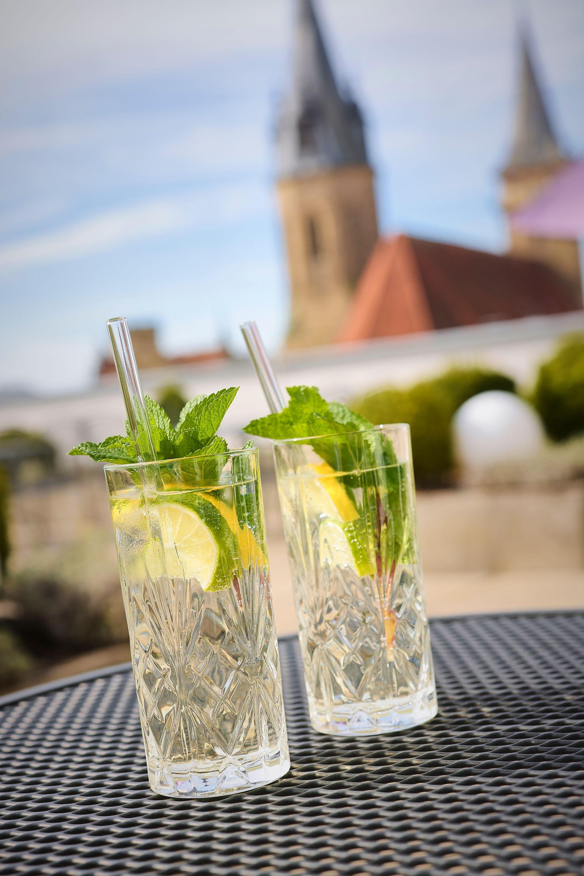 Zwei prickelnde Cocktails mit Minze und Zitrone garniert auf einer Terrasse, im Hintergrund der Kirchturm.