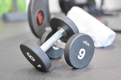 Zwei schwarze Hanteln, Gewicht 9 und 6, auf einem Fitnessraumboden. Im Hintergrund sind eine Hantelscheibe und ein Handtuch zu sehen.