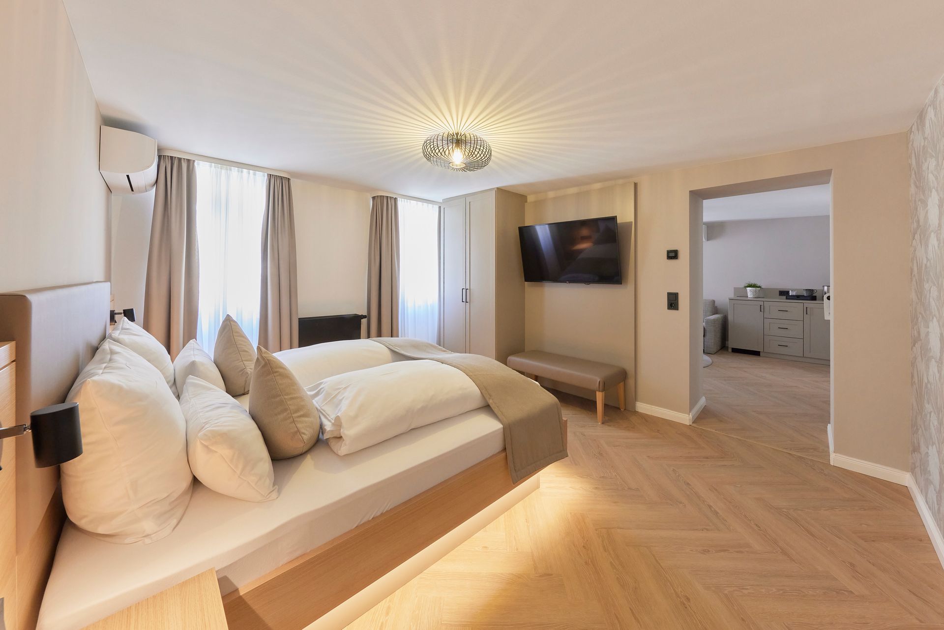 Schlafzimmer mit Bett, Fernseher, Sitzbank, Fenster und Blick in einen weiteren Raum mit Theke.