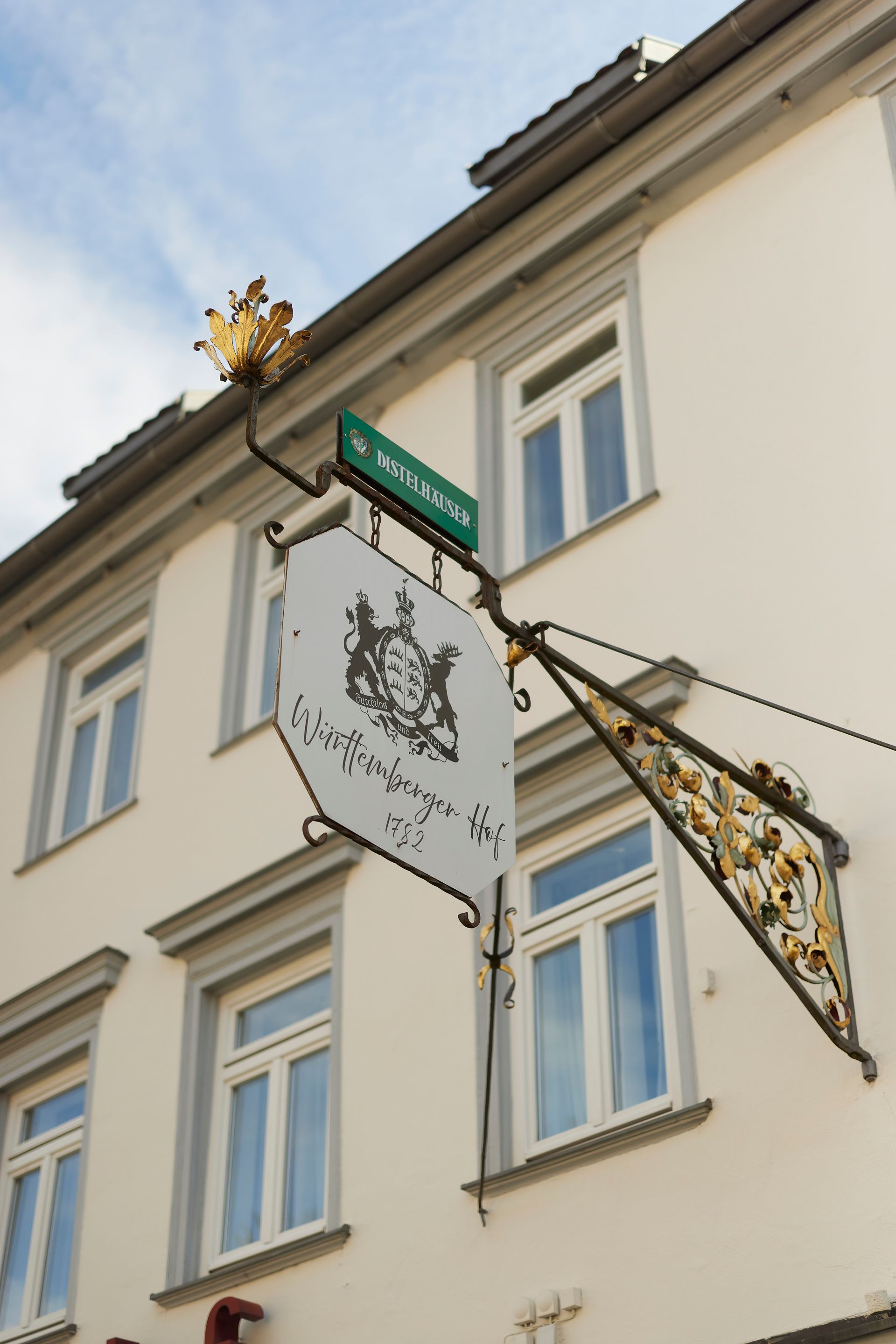 Am Gebäude hängendes Schild: verziertes Gold und Weiß, deutscher Text, Wappen.