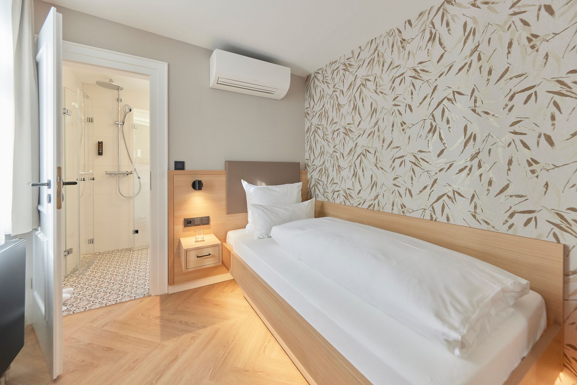 Ein Hotelzimmer mit Einzelbett, Dusche und gemusterter Wand. Farbschema in Beige und Weiß.