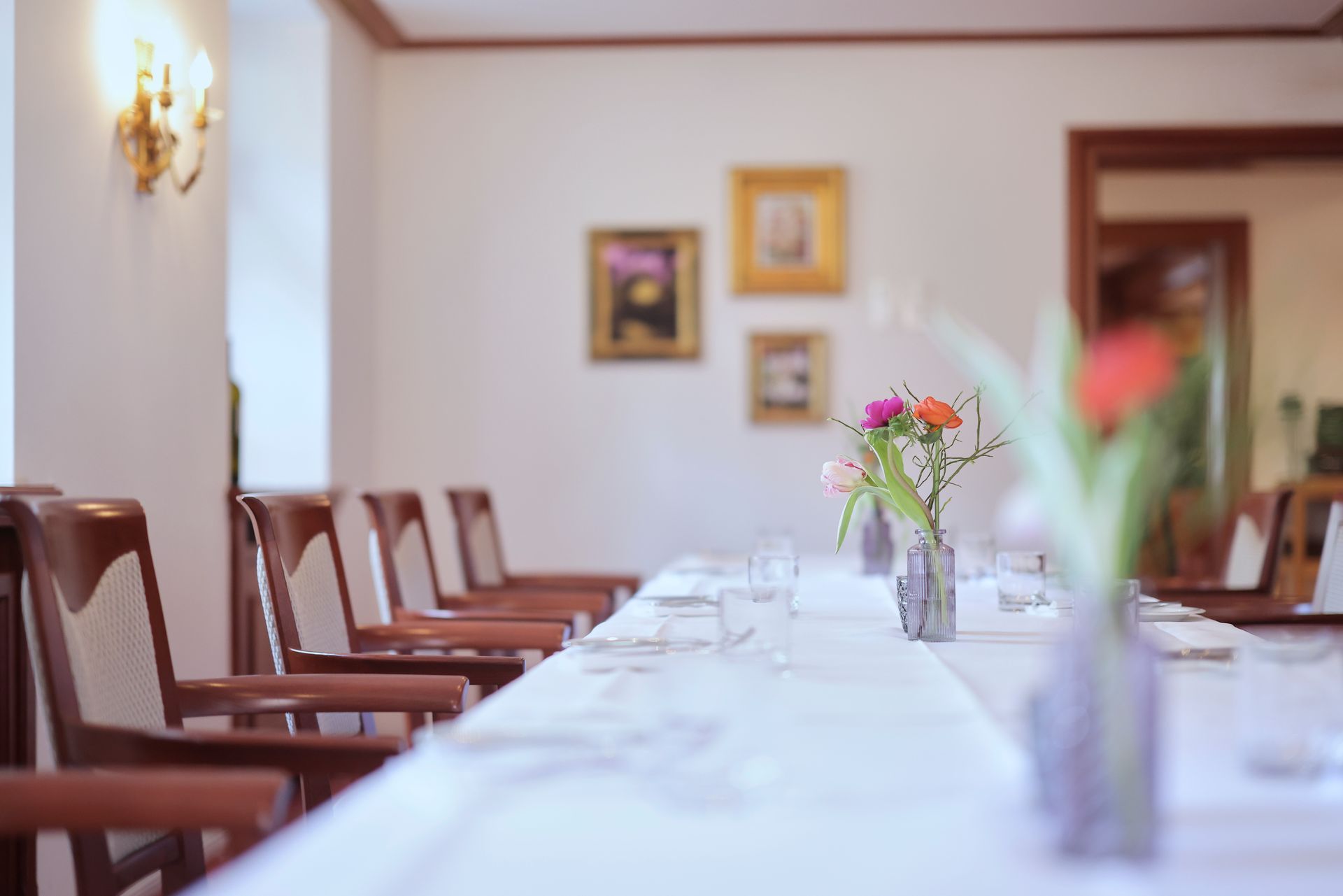 Langer Esstisch mit Gläsern und Blumen, flankiert von braunen Stühlen, mit Gemälden an der Wand.