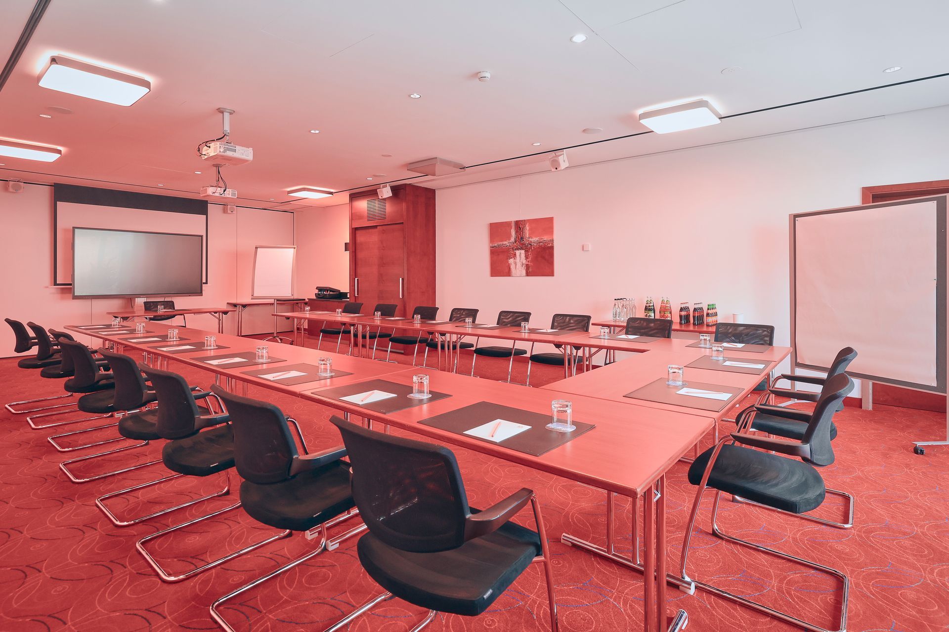 Konferenzraum mit U-förmigem Tisch und schwarzen Stühlen. Roter Teppich und Wände. Projektionsleinwand.