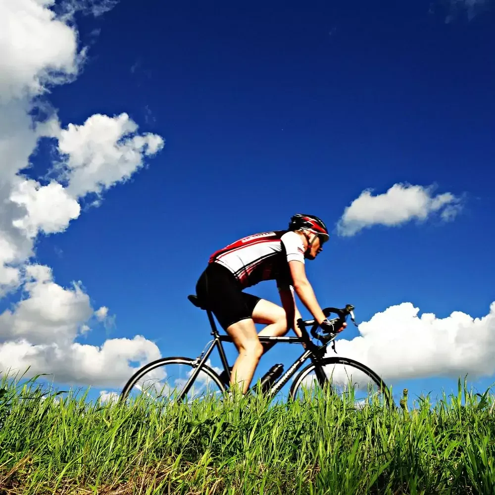 Radfahrer fährt auf Rennrad durch grünes Gras, strahlend blauer Himmel mit flauschigen weißen Wolken.