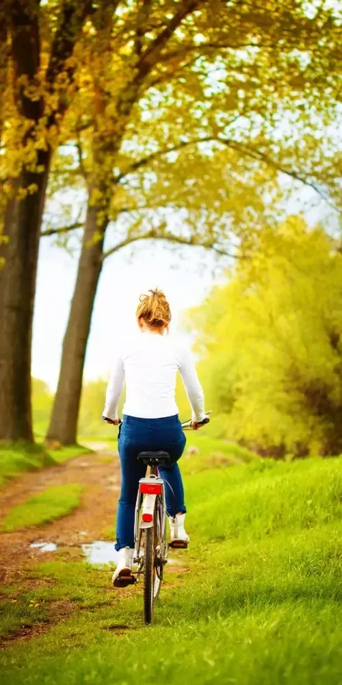 Frau fährt mit dem Fahrrad auf einem Weg durch einen sonnigen, grünen Park mit Bäumen mit goldenen Blättern.