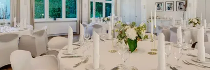 Eleganter Speisesaal, Tische für eine formelle Veranstaltung gedeckt. Weiße Tischwäsche, Blumengestecke und hohe Kerzen.