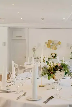 Formelles Esstischgedeck in einem hellen, weißen Raum mit floralen Tafelaufsätzen und elegantem Glasgeschirr.