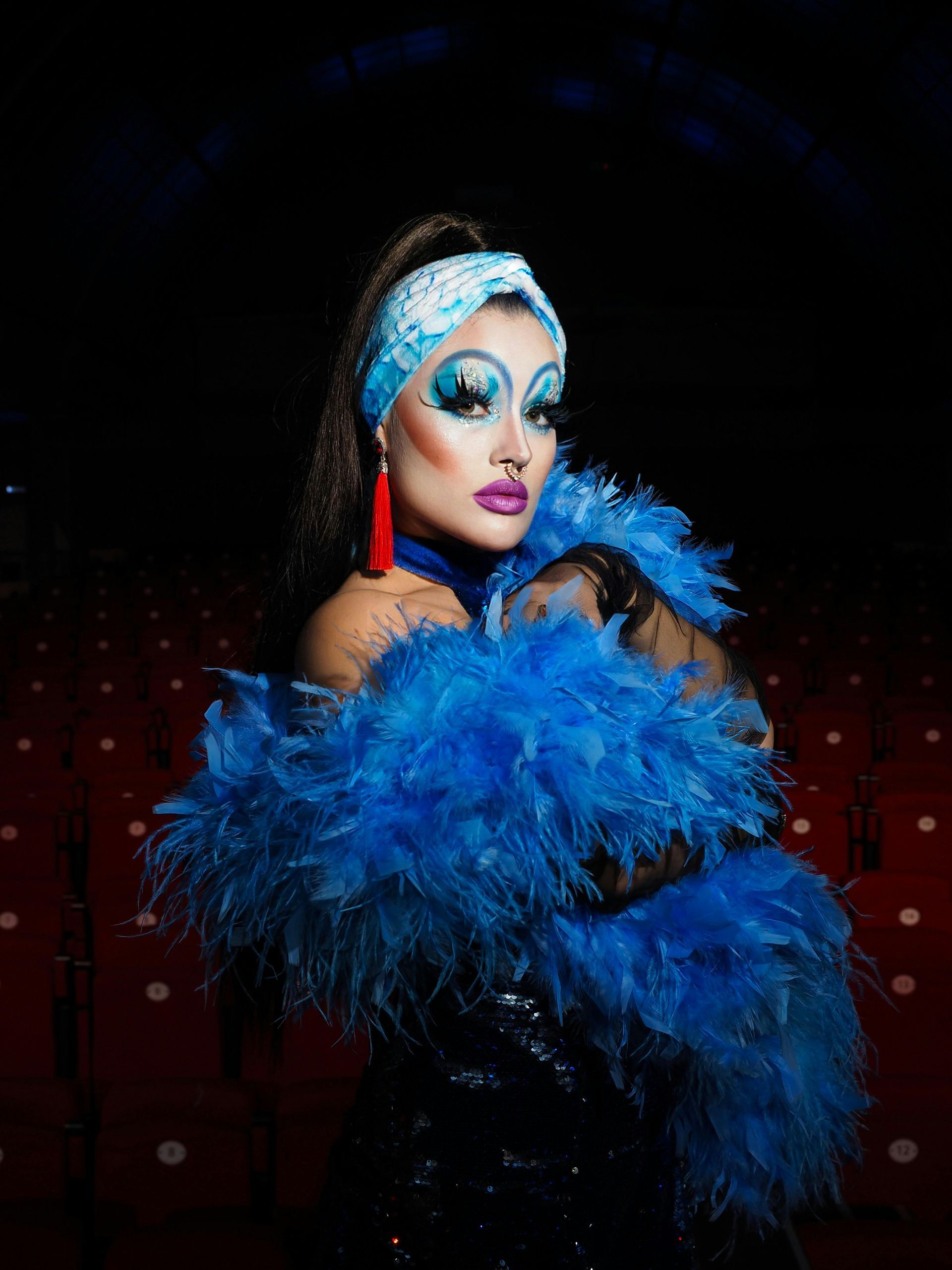 Eine Dragqueen mit blauem Make-up und Boa, die ein Stirnband und rote Ohrringe trägt und dramatisch posiert.