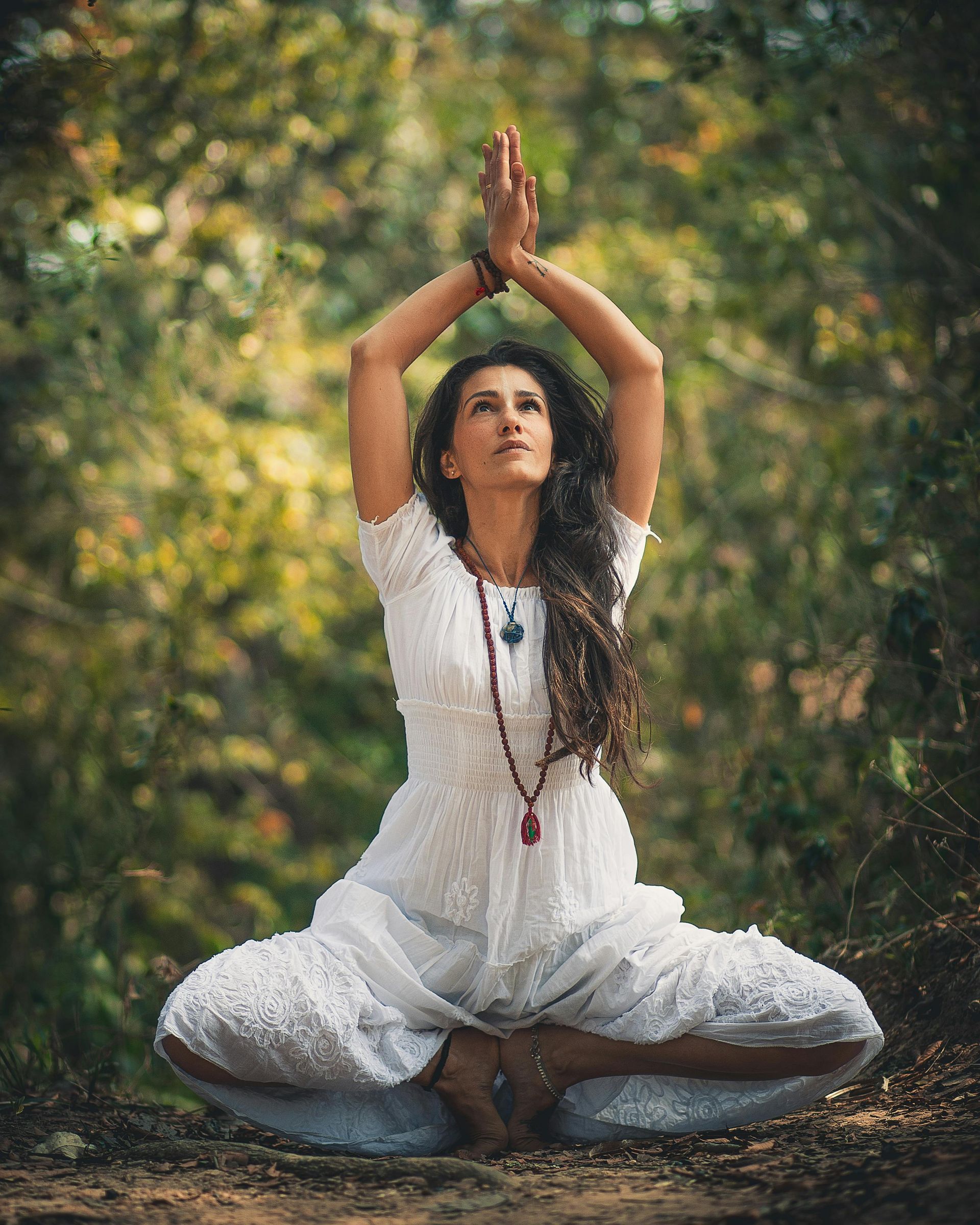 Frau im weißen Kleid, die Hände erhoben, sitzt in meditativer Pose im Freien und blickt nach oben.