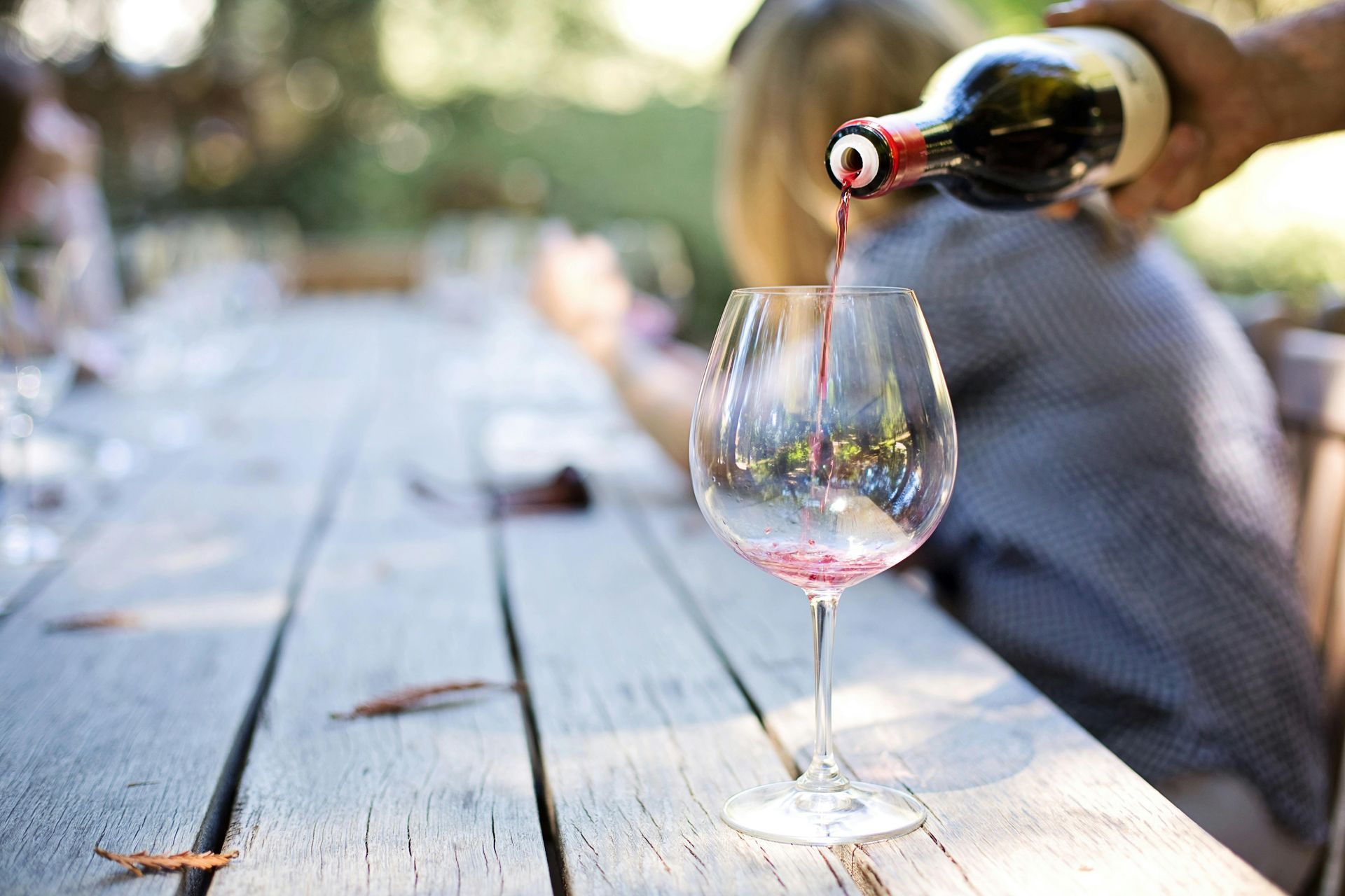 Person gießt an einem Tisch im Freien Rotwein in ein Glas, andere Personen sind im Hintergrund unscharf zu sehen.