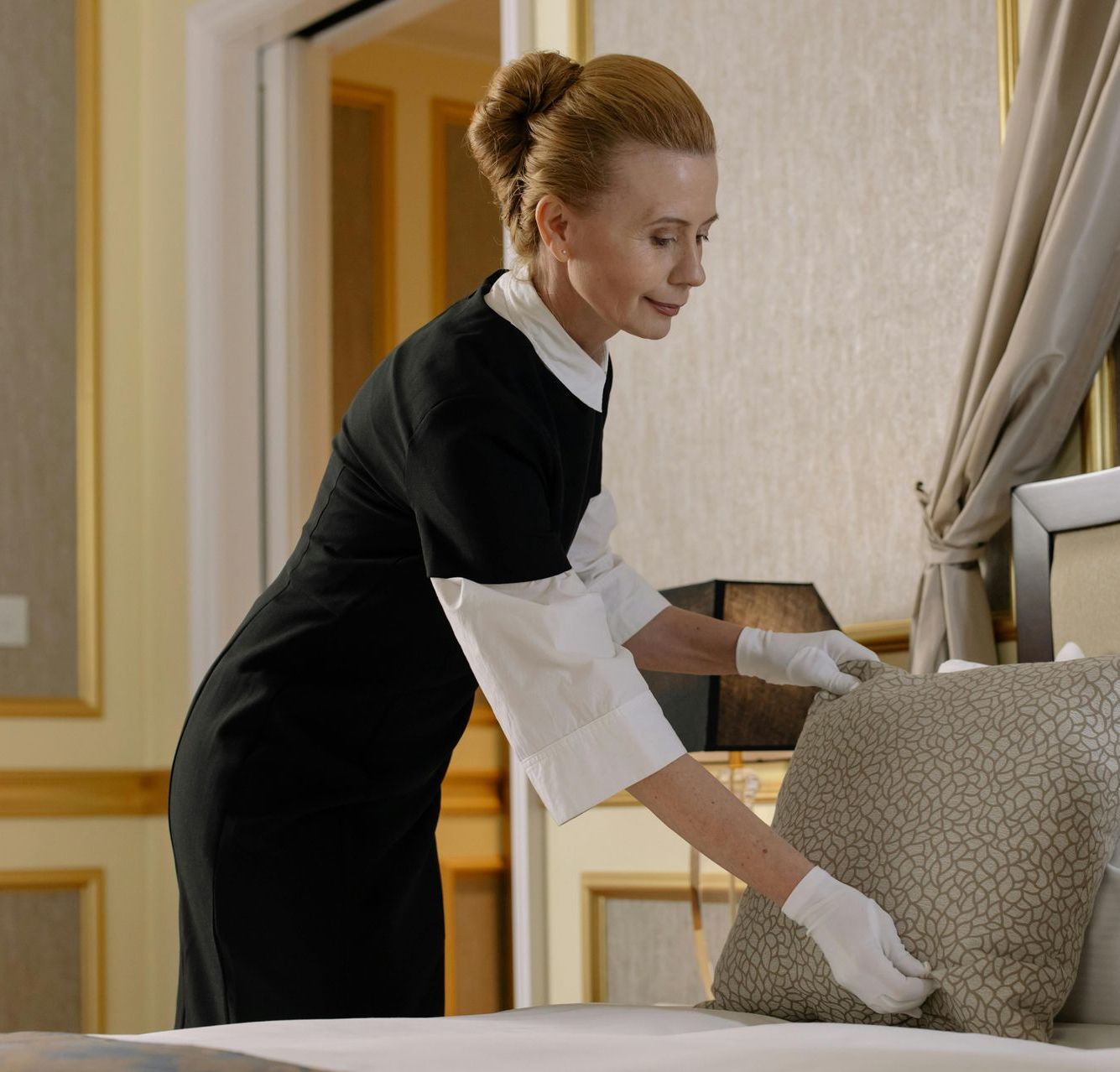 Zimmermädchen in Uniform arrangiert ein dekoratives Kissen auf einem Bett in einem Hotelzimmer.