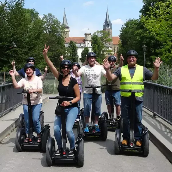 Eine Gruppe von Menschen fährt Segways auf einer Brücke und steht lächelnd und winken vor einem Schloss.