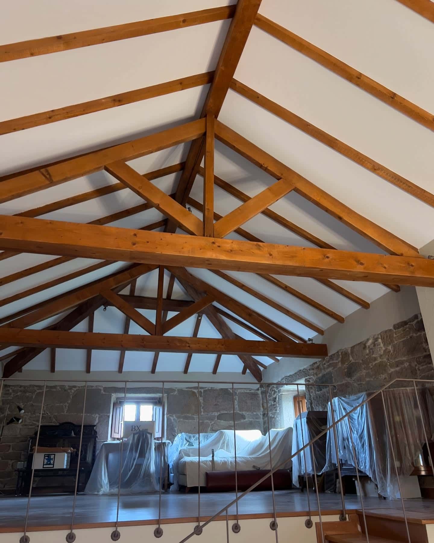 Vigas de madera sostienen un techo blanco en una habitación con una pared de piedra y muebles cubiertos.