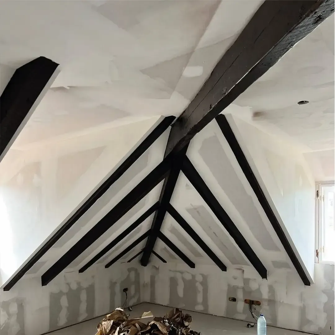 Vista interior de un ático con vigas de madera oscura expuestas contra paredes de yeso sin terminar.