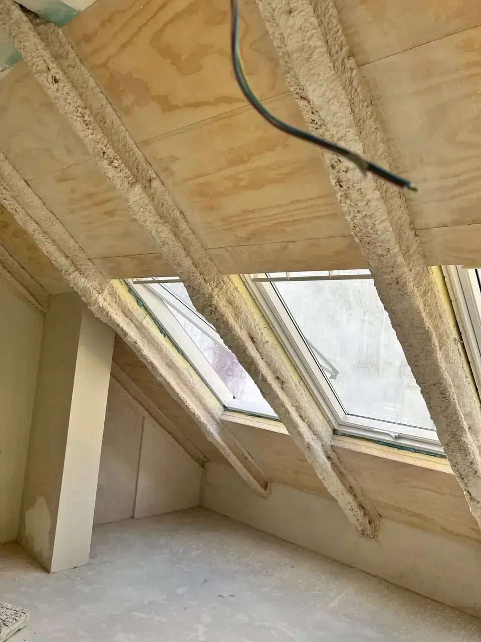 Vista interior de un ático en construcción con vigas de madera, techo de madera contrachapada y tragaluces.