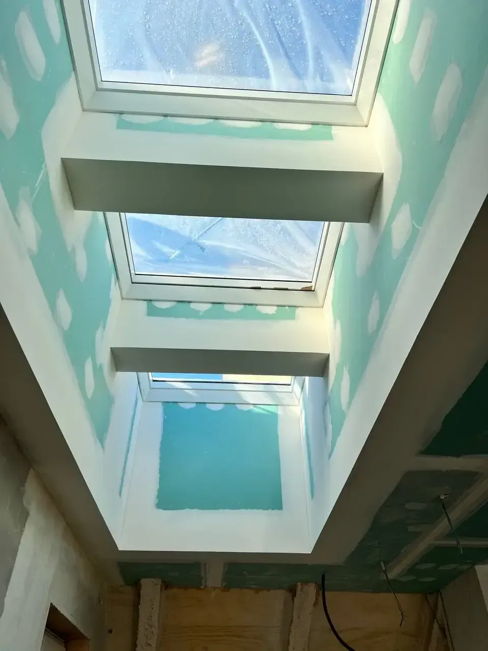 Vista interior de una habitación con tres tragaluces, con paneles de yeso y marcos expuestos; tonos azul verdoso y blanco.