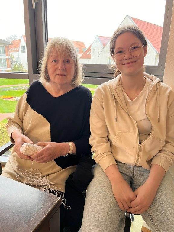 Zwei Personen sitzen an einem Fenster: eine ältere Frau in einem schwarz-cremefarbenen Oberteil, die einen Gegenstand hält; eine jüngere Person in einem hellen Kapuzenpullover, die lächelt.