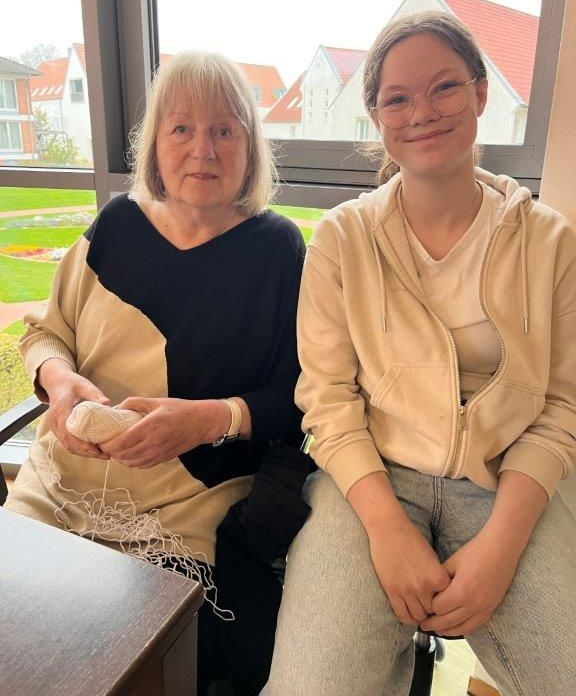 Zwei Personen sitzen an einem Fenster: Ältere Frau hält einen Gegenstand, jüngere Person lächelt, helle Kleidung, neutrale Umgebung.
