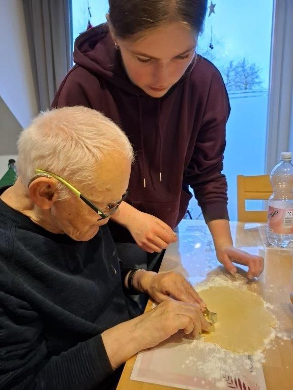 Ein junger Mensch hilft einem älteren Menschen dabei, Kekse aus Teig auf einem Tisch auszustechen.