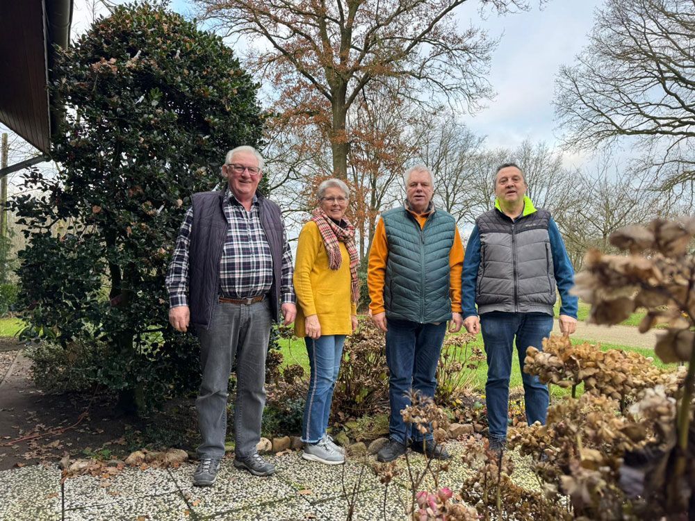 Vier Personen stehen im Freien. Hinter ihnen befinden sich ein grüner Busch und ein Baum. Der Himmel ist bedeckt.