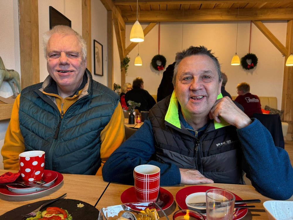 Zwei Männer sitzen lächelnd an einem Tisch in einem Café. Sie sitzen vor Tassen und Tellern.