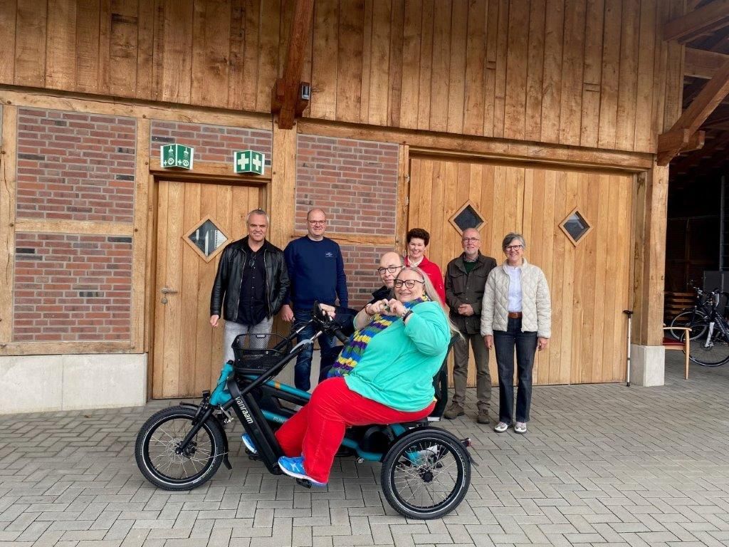 Eine Gruppe posiert vor einem Holzgebäude mit einem türkisfarbenen Dreirad. Die Person in roter Hose und türkisfarbenem Oberteil sitzt auf dem Dreirad.