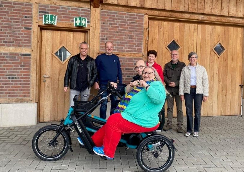 Eine Gruppe von Menschen posiert vor einem Holzgebäude mit einem türkisfarbenen Dreirad. Der Fahrer des Dreirads zeigt den Daumen nach oben.