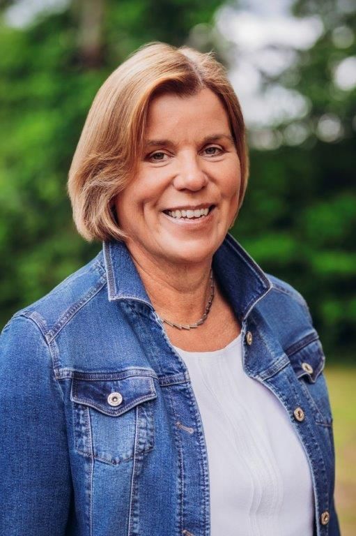 Frau lächelt, trägt eine Jeansjacke und steht im Freien vor einem verschwommenen grünen Hintergrund.