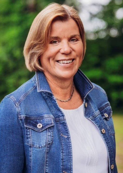 Frau lächelt, sie trägt eine blaue Jeansjacke über einem weißen Hemd, vor einem verschwommenen grünen Hintergrund.