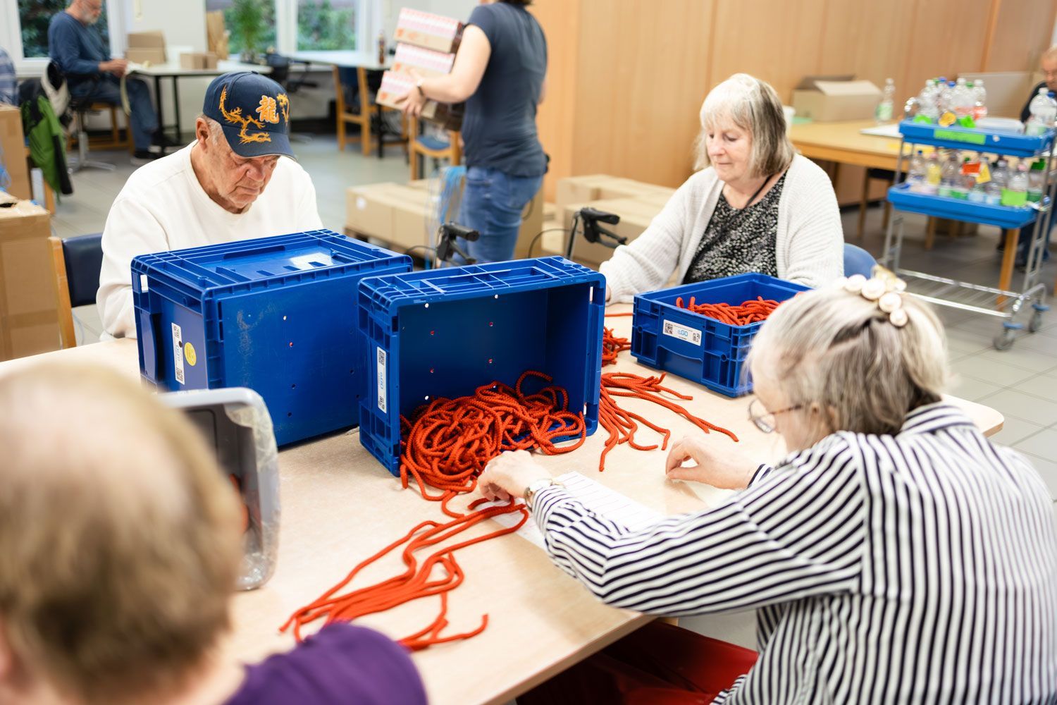 Menschen sortieren rote Kabel an einem Tisch mit blauen Kisten; jemand trägt eine Kiste.