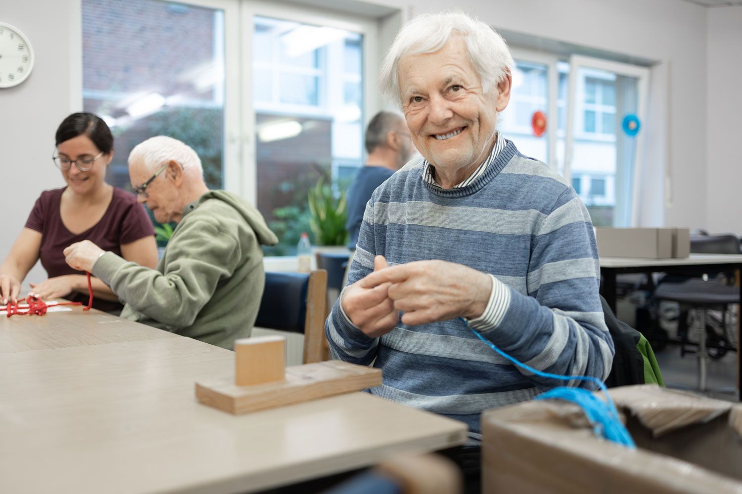 Ein lächelnder älterer Mann strickt in einem hell erleuchteten Raum; andere basteln an einem Tisch mit einer Betreuerin.
