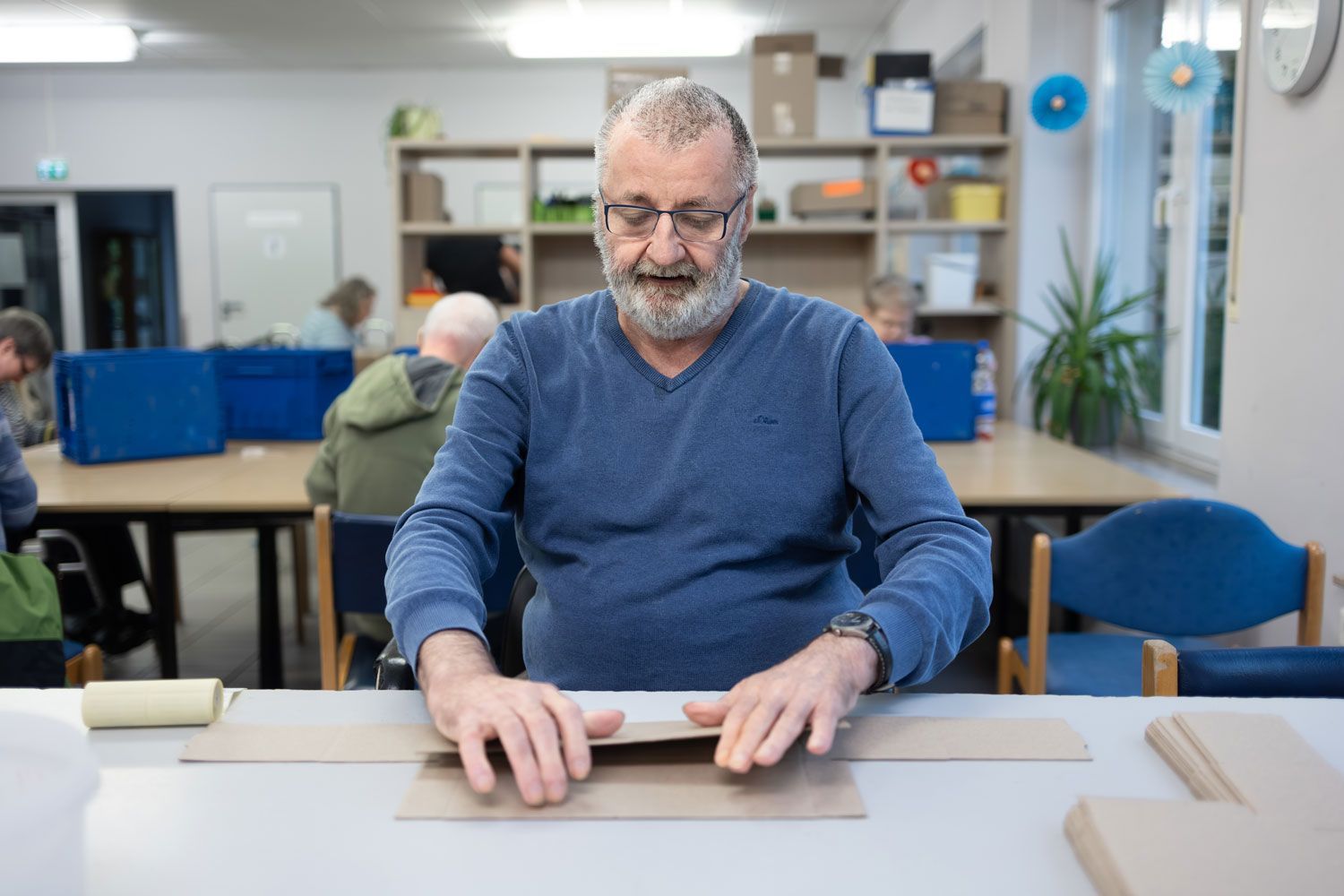 Ein Mann mit Brille faltet Pappe an einem Tisch in einer Werkstatt; im Hintergrund sind andere Personen zu sehen.