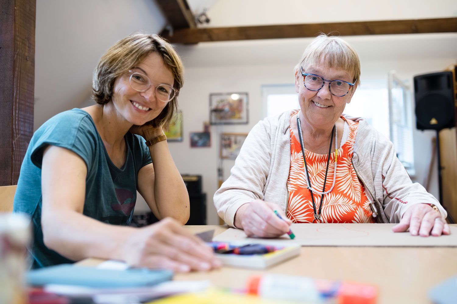 Zwei Personen sitzen lächelnd an einem Tisch. Eine zeichnet mit einem grünen Stift. Sie befinden sich in einem Raum mit Kunstwerken an den Wänden.