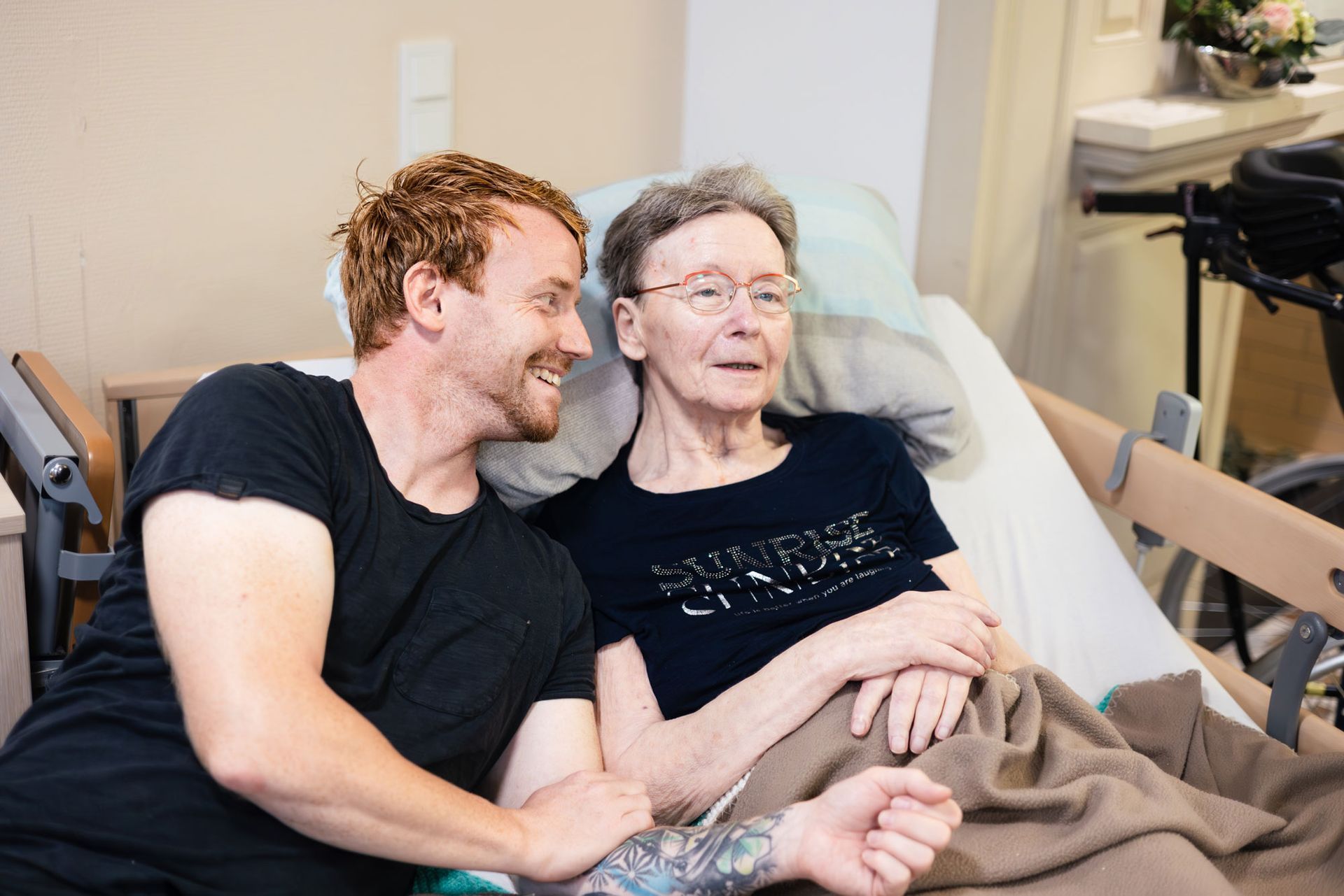 Junge Frau umarmt ältere Frau im Rollstuhl; beide lächeln, im Freien.