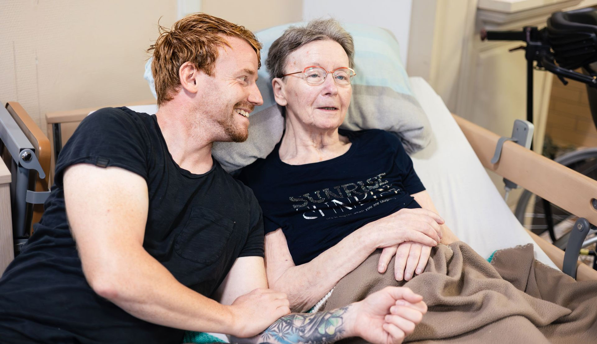 Pflegekraft führt ältere Frau im Rollstuhl durch einen Blumengarten.