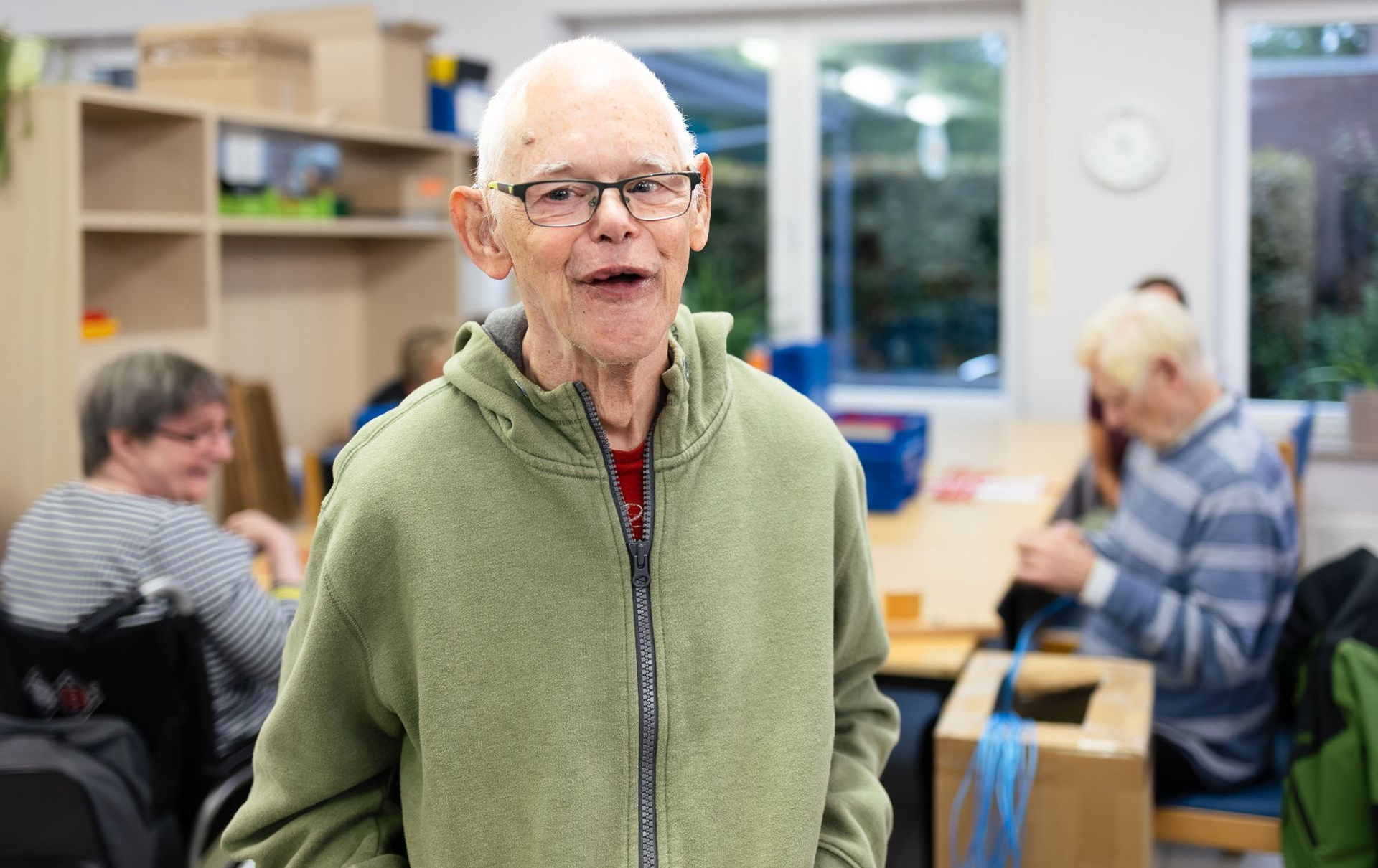 Mann im gelben Hemd lächelt und gestikuliert zum klatschenden Mann. Gruppe sitzt draußen.