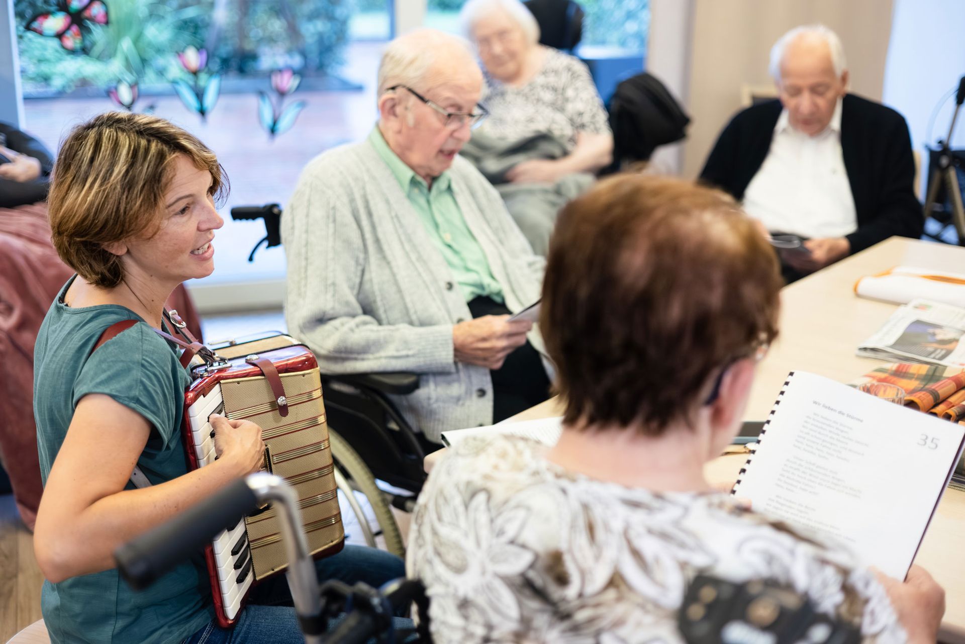 Eine Pflegekraft liest einem älteren Paar im Freien vor. Der Mann benutzt einen Rollator, die Frau einen Rollstuhl.