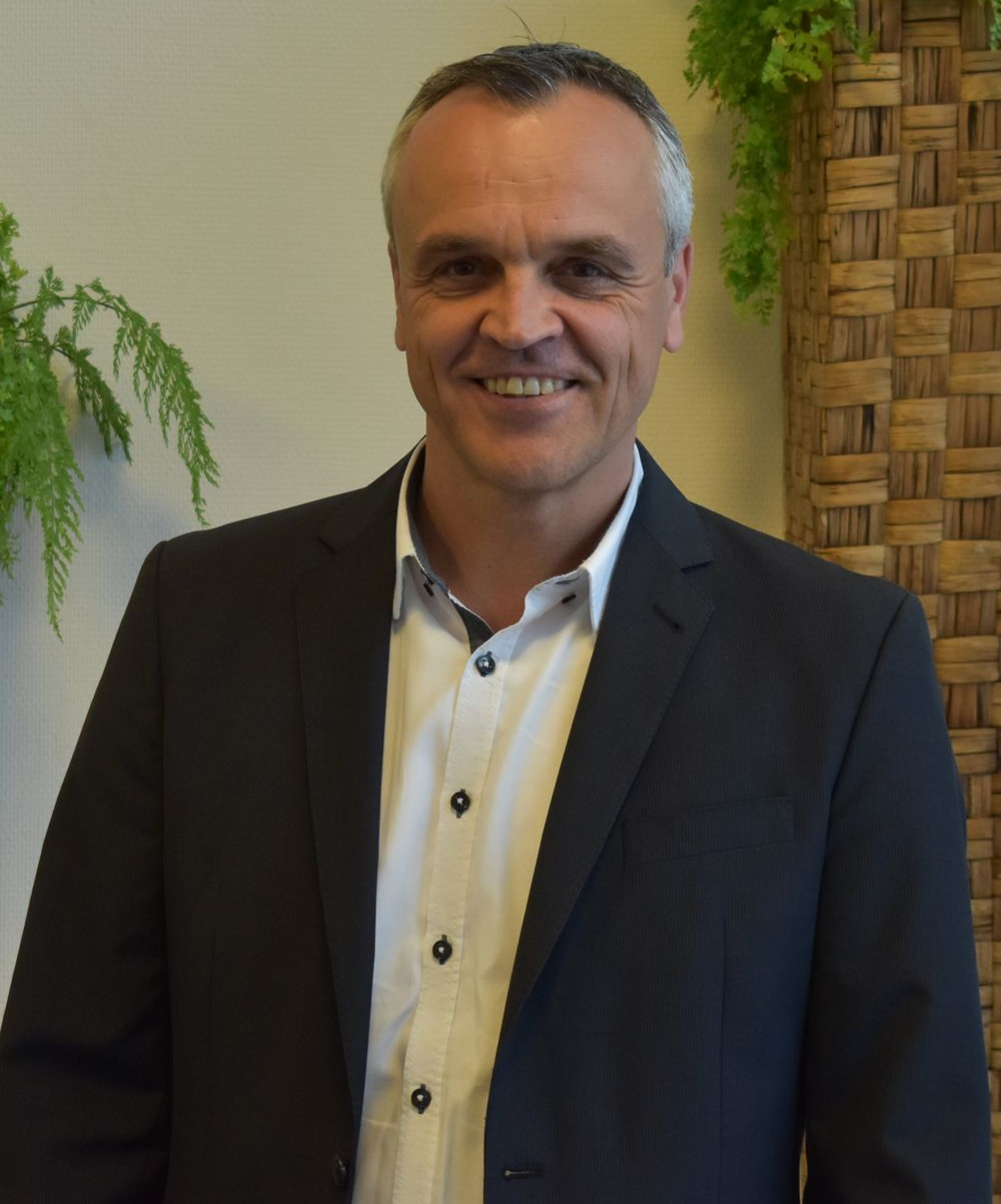 Mann im Anzug lächelt, steht drinnen vor einer geflochtenen Wand und Pflanzen.