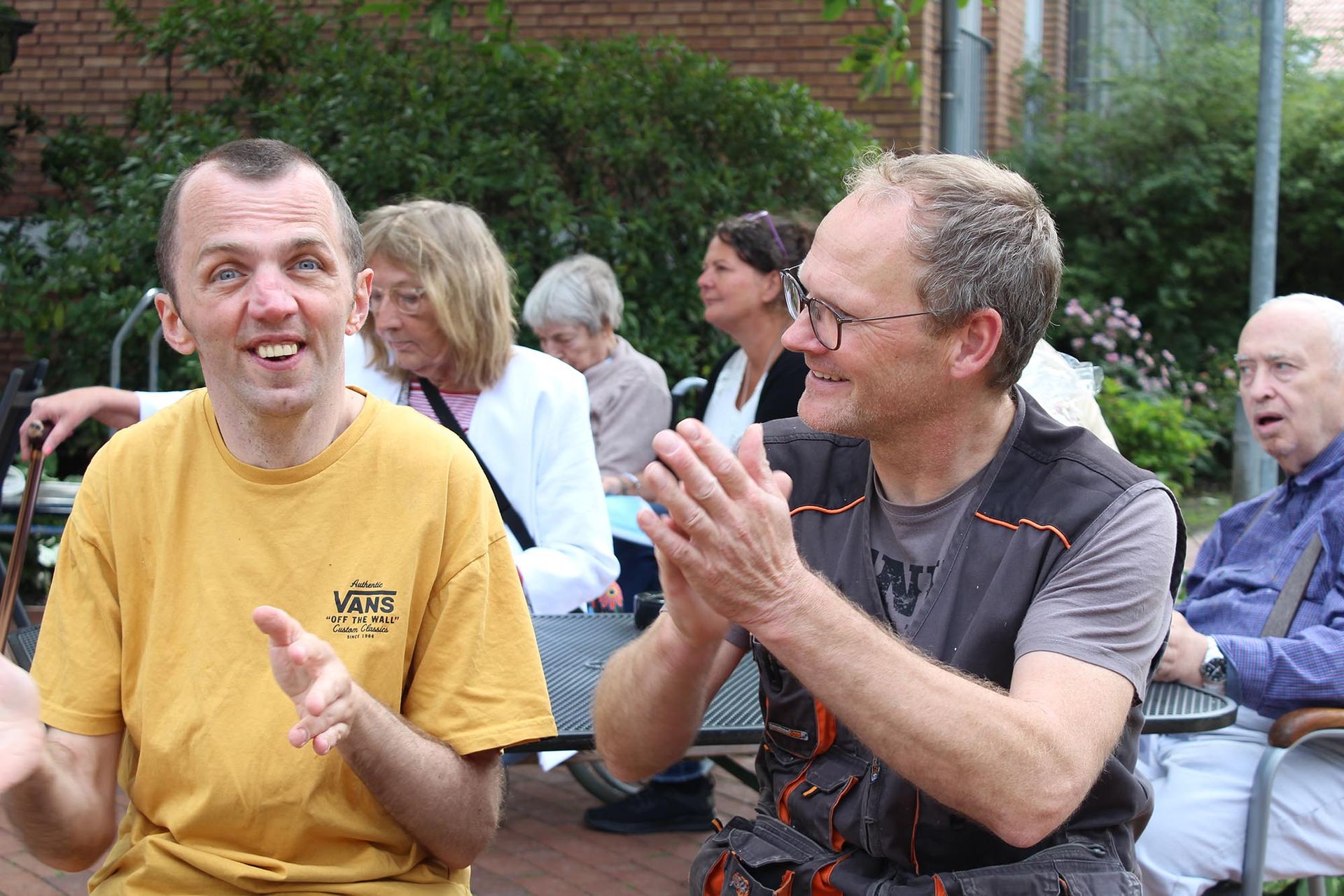 Mann im gelben Hemd lächelt und gestikuliert. Mann klatscht, sitzt mit Gruppe im Freien.