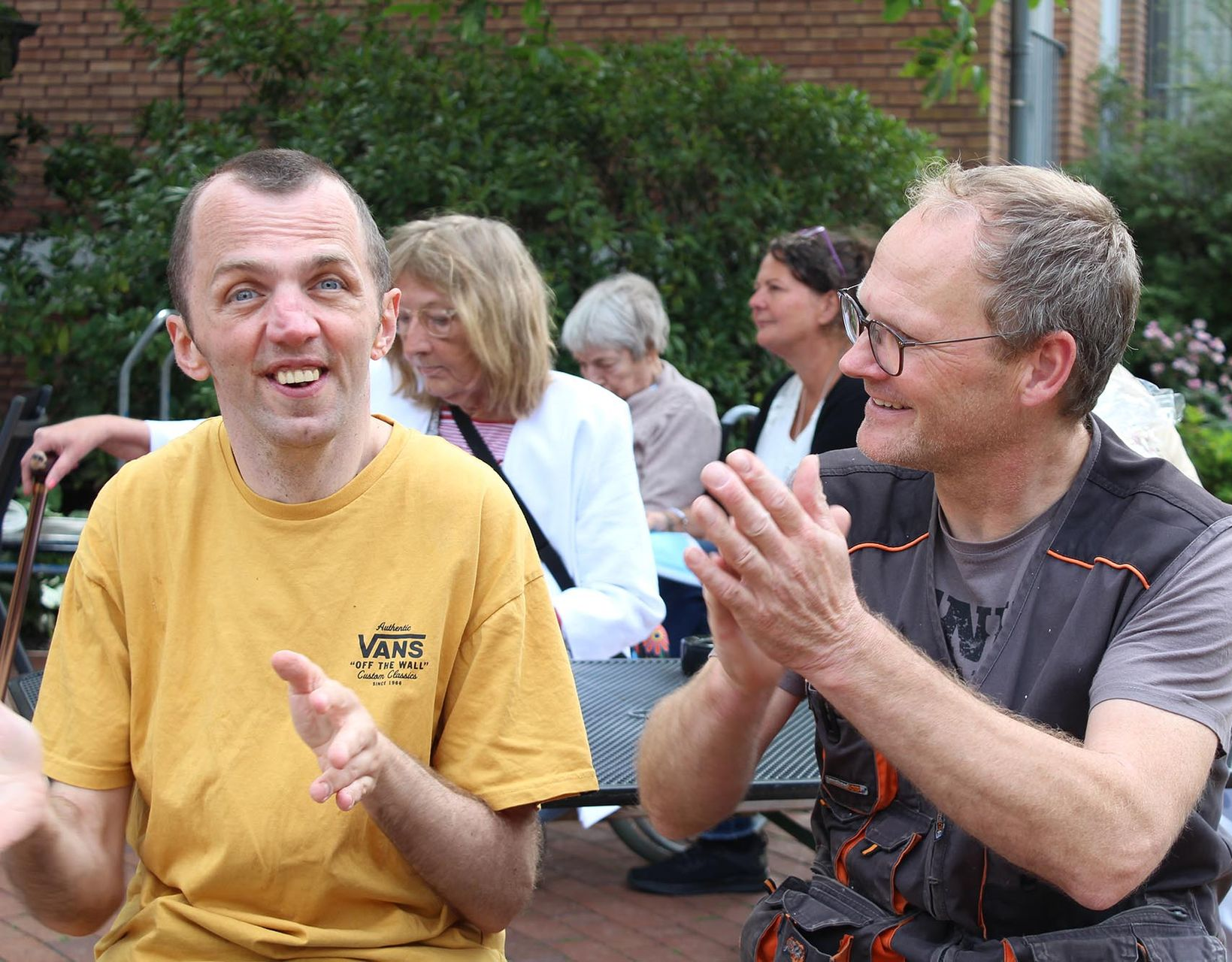 Zwei Männer, einer im gelben Hemd, lächelnd, der andere klatschend. Mehrere Personen im Hintergrund. Außenaufnahme.
