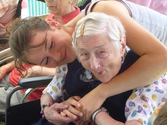 Junge Frau umarmt ältere Frau im Rollstuhl; beide lächeln, im Freien.