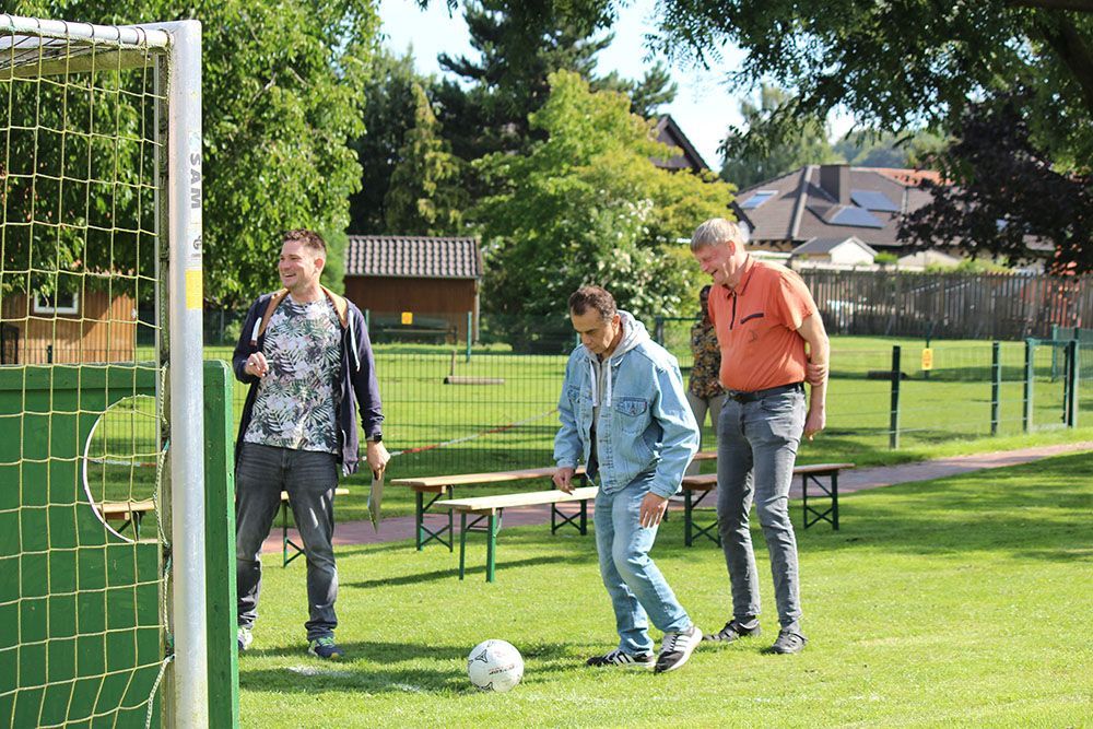 Drei Männer auf einer Wiese mit einem Fußball. Einer kickt den Ball, zwei schauen zu. Grüner Torpfosten links.
